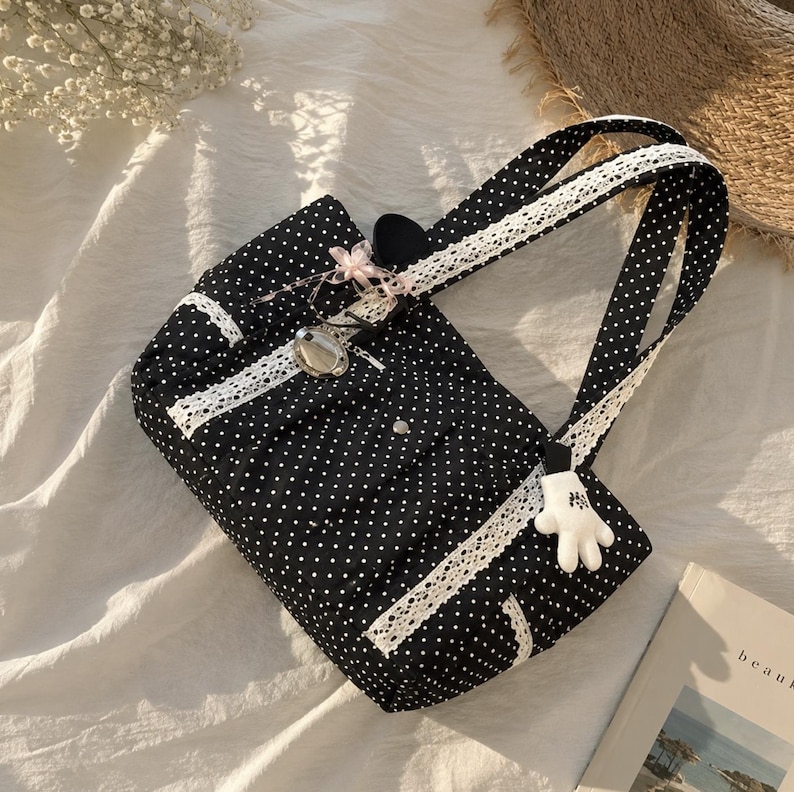 Puede incluir: Un bolso tote negro con estampado de lunares blancos, con ribete de encaje blanco a lo largo de las asas y el panel frontal. El bolso incluye un peque&ntilde;o dije plateado, un detalle de cinta rosa y un llavero con forma de guante blanco.