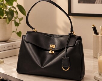 Puede incluir: Un bolso de mano de cuero negro con una sola asa superior, una solapa con cierre de giro de metal dorado y una etiqueta de cuero a juego con un anillo dorado sujeto al lateral mediante una fina correa de cuero.