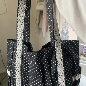 Puede incluir: Un bolso tote negro con un patr&oacute;n de lunares blancos en toda la superficie. El bolso incluye dos correas para el hombro y bolsillos laterales, cada uno acentuado con una tira vertical de encaje blanco. El dise&ntilde;o es uniforme en el cuerpo y las correas.