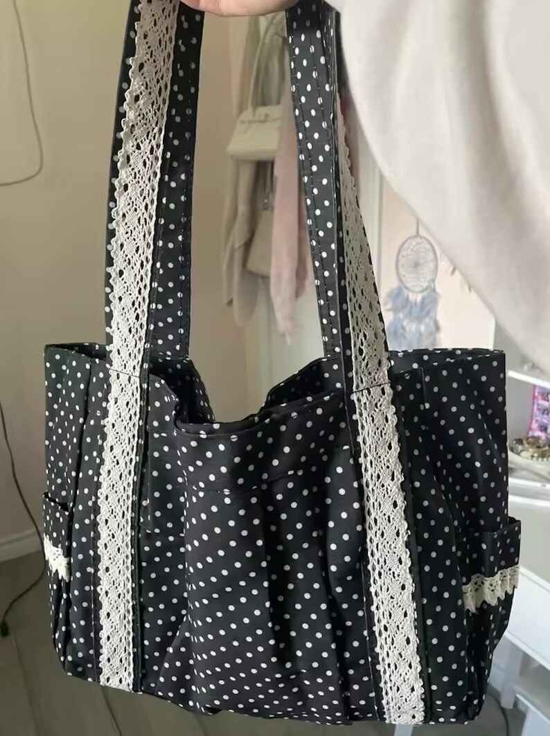 Puede incluir: Un bolso tote negro con un patr&oacute;n de lunares blancos en toda la superficie. El bolso incluye dos correas para el hombro y bolsillos laterales, cada uno acentuado con una tira vertical de encaje blanco. El dise&ntilde;o es uniforme en el cuerpo y las correas.