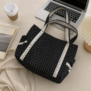Puede incluir: Un bolso tote negro con un patr&oacute;n de lunares blancos y ribete de encaje blanco a lo largo de las dos asas y el panel frontal. El bolso tiene una estructura rectangular con dos asas superiores y una confecci&oacute;n en tela con dise&ntilde;o de puntos.