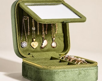 Puede incluir: Un joyero cuadrado de terciopelo verde oliva con cremallera dorada. La tapa abierta cuenta con un espejo rectangular y cuatro collares dorados colgados de ganchos. La base contiene cuatro ranuras acolchadas para anillos.