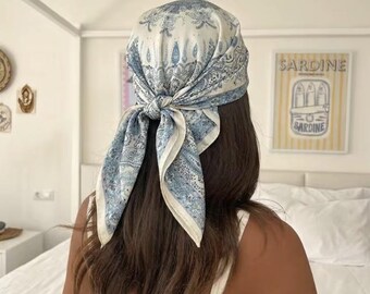 Puede incluir: Un pañuelo cuadrado atado en la parte posterior de la cabeza, con un detallado estampado de cachemira en azul y blanco. La tela muestra intrincados motivos florales y ornamentales en toda la superficie, con un centro anudado y dos extremos sueltos.