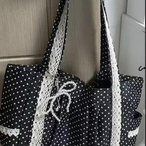 Puede incluir: Un bolso tote negro con estampado de lunares blancos, acentuado con ribetes de encaje blanco vertical en la parte delantera y las correas. Un dije decorativo de lazo de perlas artificiales blancas cuelga del frente y los bolsillos laterales tienen encaje blanco.