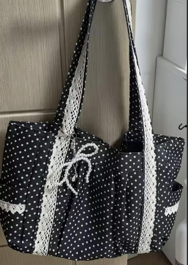 Puede incluir: Un bolso tote negro con estampado de lunares blancos, acentuado con ribetes de encaje blanco vertical en la parte delantera y las correas. Un dije decorativo de lazo de perlas artificiales blancas cuelga del frente y los bolsillos laterales tienen encaje blanco.