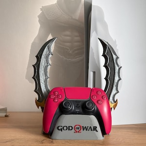 Puede incluir: Un mando de videojuegos rosa y negro sobre un soporte gris con el texto GOD OF WAR. Detrás del mando hay dos hojas curvas de aspecto metálico con detalles dorados, situadas delante de una consola de videojuegos blanca.