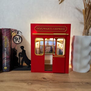 May include: A red Hogwarts Express train carriage book nook insert featuring gold lettering, a sliding door, and interior lighting. The structure displays a rectangular sign with the text HOGWARTS EXPRESS and a detailed view of a train cabin interior.