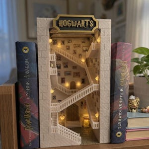Könnte beinhalten: Ein beigefarbener Buchstützen-Einsatz mit einem Miniatur-Innenraum einer Schlosstreppe mit weißen Geländern, mehreren kleinen gerahmten Bildern an den Wänden und warmer Innenbeleuchtung. Ein schwarzes Schild mit goldener Aufschrift HOGWARTS.
