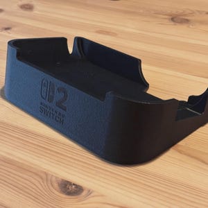 Può includere: Una docking station nera in plastica testurizzata per console Nintendo Switch, con il logo Nintendo Switch in rilievo sul lato anteriore. Il supporto presenta una base rettangolare con ritagli sagomati per alloggiare la console.