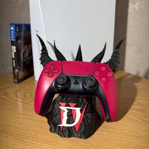 May include: A bright pink wireless video game controller rests on a black, textured display stand featuring a white Roman numeral IV logo. The stand includes black, pointed, wing-like protrusions extending upward behind the controller.