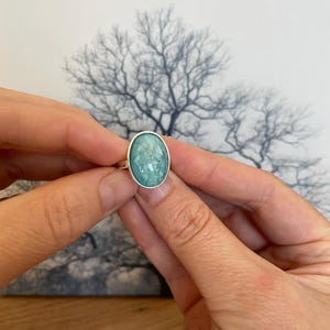 Puede incluir: Un anillo de plata con una piedra ovalada pulida de color azul verdoso con inclusiones blancas veteadas. La piedra preciosa está engastada en un bisel de plata liso y sencillo, mostrando un patrón natural variado en su superficie.