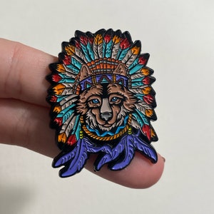 May include: A colourful enamel pin featuring a wolf wearing a Native American headdress with feathers in shades of red, orange, blue, and green.