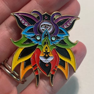 May include: A colorful enamel pin with a black background and a rainbow design. The pin features a stylized face with a third eye and a flower-like design.