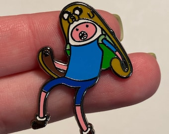 Puede incluir: Un personaje de dibujos animados con un sombrero amarillo y una camisa azul sostiene una mochila marrón. El personaje está en una pose de carrera con la boca abierta. El pin es de metal y tiene un acabado brillante.