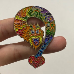May include: A colorful enamel pin shaped like a question mark with a dragon head in the middle. The pin is decorated with a rainbow of colors and the words "Mystery Land" are on the bottom.