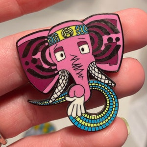 Puede incluir: Un pin de elefante rosa con una diadema colorida y un patrón de remolino en sus orejas. La trompa del elefante está enroscada en un círculo y tiene un patrón azul, amarillo y verde.