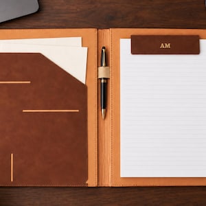 Può includere: Una cartella portadocumenti in pelle color cuoio e marrone scuro con una tasca sinistra contenente carta bianca, un passante centrale per penna con una penna nera e oro e un blocco note destro con intestazione marrone scuro e le lettere AM.