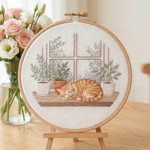 Könnte beinhalten: Ein Stickrahmen mit einer gestickten Szene einer orangefarbenen getigerten Katze, die auf einem Fensterbrett schläft. Das Design umfasst drei Topfpflanzen und einen Fensterrahmen auf weißem Stoff. Der Rahmen ist mit einer Messingschraube fixiert.