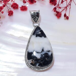 Puede incluir: Un colgante en forma de lágrima con una piedra pulida en blanco y negro engastada en un marco decorativo de plata de ley. El pasador de plata en la parte superior muestra un intrincado diseño de volutas que sujeta la piedra.