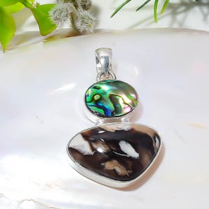 May include: A silver pendant featuring a top oval-shaped iridescent abalone shell inlay with green and blue tones, connected to a larger bottom stone with a smooth, irregular shape displaying a marbled pattern of dark brown, black, and white colors.