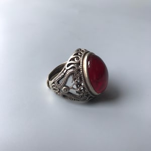 Puede incluir: Un anillo de metal plateado con una piedra roja translúcida, lisa y de forma ovalada en el centro. La banda del anillo presenta intrincados detalles de filigrana calada que envuelven los lados, creando una apariencia texturizada y decorativa.