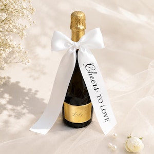 Peut inclure: Une petite bouteille de champagne avec un col en aluminium doré et une étiquette dorée portant le mot "Lucky" en écriture cursive. Un ruban de satin blanc est noué autour du col, avec le texte "Cheers TO LOVE" imprimé en noir sur un pan.