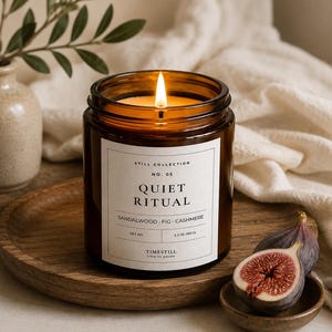Puede incluir: Una vela encendida en un frasco de vidrio ámbar con una etiqueta blanca que dice Still Collection No. 05 Quiet Ritual Sandalwood Fig Cashmere Net Wt 5.3 oz (150 g) Timestill time to pause. La vela descansa sobre una bandeja redonda de madera.