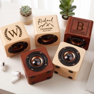 May include: Five cube-shaped wooden speakers with circular black and copper-coloured drivers. Three speakers feature custom laser-engraved designs: a letter W with a laurel wreath, the name Richard with mountains, and the letter B with the name Bernard.
