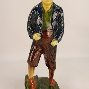 Könnte beinhalten: Eine glasierte Keramikfigur einer Person mit rotbraunem Haar, bekleidet mit einer dunkelblauen Jacke über einem grünen Hemd, brauner Hose und rotbraunen Schuhen. Die Figur steht auf einem rechteckigen, glänzend grünen Sockel.