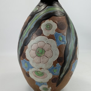 May include: A ceramic vase with a brown base color, featuring a dark brown neck and rim. The body is decorated with white floral patterns, light green accents, and blue geometric shapes, all outlined in dark brown, creating a textured appearance.