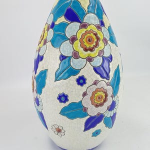 Op de afbeelding: Een keramische vaas met een crèmekleurige craquelé glazuurafwerking, voorzien van een levendig bloemmotief in tinten blauw, geel, paars en wit. De vaas heeft een smalle hals en een ronde vorm, met een effen blauwe rand en onderkant.