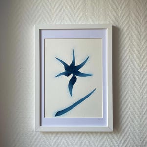 Peut inclure: Une œuvre d'art encadrée représentant une forme florale abstraite bleu foncé minimaliste avec six pétales pointus et une tige incurvée en dessous, centrée sur un fond crème dans un cadre rectangulaire blanc simple.