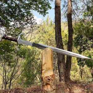 May include: A long sword with a silver-toned metal blade featuring a textured dark section near the hilt, a cross-shaped guard, and a dark brown wooden handle with metallic rivets, positioned horizontally atop a vertical wooden log.
