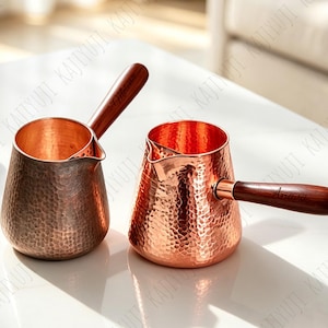 以下が含まれることがあります： 暗い木製のハンドルが付いた2つのハンマー仕上げの銅製コーヒーポット。各ポットは広い底、先細りの本体、小さな注ぎ口が特徴です。金属の表面は質感のあるくぼんだ仕上げで、ハンドルは側面に付いています。
