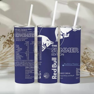 Könnte beinhalten: Drei zylindrische Becher in Dunkelblau mit weißem Text und Grafiken. Jeder verfügt über das Red Bull Summer Edition Logo, Nährwertangaben und einen weißen Strohhalm. Das Design enthält den Text The Summer Edition, Blueberry Flavour und Red Bull.