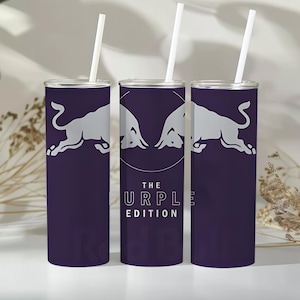 Könnte beinhalten: Drei zylindrische violette Becher mit weißen Strohhalmen. Jeder zeigt eine silberne Grafik von zwei sich gegenüberstehenden Stieren. Der mittlere Becher zeigt den Text THE PURPLE EDITION in weißer Schrift, zentriert unter der Grafik.