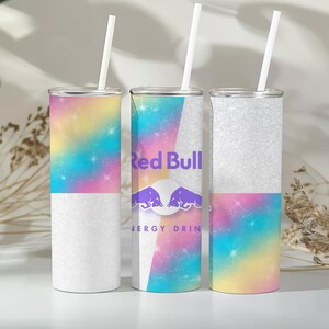 Könnte beinhalten: Drei zylindrische Trinkbecher mit weißen Strohhalmen. Jeder Becher hat ein geteiltes Design mit einem glitzernden silbernen Bereich und einem bunten Regenbogen-Nebel-Muster mit Sternen. Der mittlere Becher zeigt das Red Bull Logo.