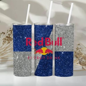 Op de afbeelding: Drie cilindervormige bekers met witte rietjes, voorzien van een glinsterende blauwe en zilveren afwerking. De middelste beker toont het rode Red Bull logo en tekst, terwijl de andere bekers afwisselende blauwe en zilveren glitters hebben.