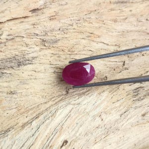 May include: A deep red, oval-cut gemstone held by a pair of metal tweezers. The stone features a faceted surface that reflects light, showcasing a vibrant crimson hue. The gemstone is positioned centrally, displaying its polished and symmetrical shape.