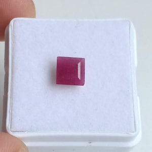 Puede incluir: Una piedra preciosa de color rojo rosado intenso, tallada en forma cuadrada con una superficie facetada, centrada sobre una almohadilla de exhibición blanca. La piedra muestra una calidad translúcida con una forma geométrica definida.