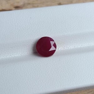 Op de afbeelding: Een ronde, gefacetteerde, dieprode robijn edelsteen. De steen heeft een briljant slijpvorm met meerdere driehoekige facetten die het licht reflecteren, waardoor een helder geometrisch patroon op het oppervlak ontstaat. De robijn ligt op een wit, gestructureerd oppervlak.