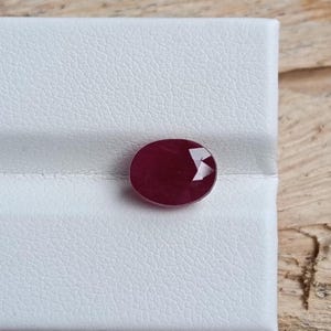 Puede incluir: Una piedra preciosa de color rojo intenso y forma ovalada con una superficie facetada. La piedra muestra un tono carmesí rico y saturado, colocada sobre un soporte de exhibición blanco texturizado. Presenta múltiples facetas talladas.