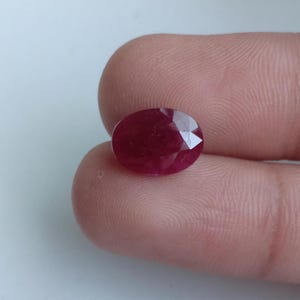 Może przedstawiać: Owalny, fasetowany kamień szlachetny rubin o głębokim czerwonym kolorze. Kamień posiada polerowaną powierzchnię z wieloma trójkątnymi fasetami, które odbijają światło, ukazując jego półprzezroczystą jakość oraz bogaty, nasycony karmazynowy odcień.