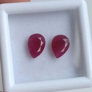 May include: Two pear-shaped, faceted rubies with a deep red color are displayed side-by-side within a square white plastic display case. The gemstones exhibit a polished surface with visible internal facets that reflect light across their surfaces.