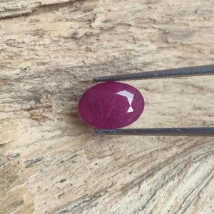 May include: An oval-shaped, deep magenta-coloured gemstone with a faceted surface held by a pair of metal tweezers. The stone displays a translucent quality with visible internal inclusions and a polished finish, reflecting light on its top facets.