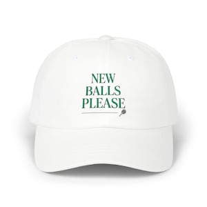 Pode incluir: Um boné de beisebol branco com o texto verde escuro NEW BALLS PLEASE impresso numa fonte serif na parte da frente. Abaixo do texto encontra-se uma linha horizontal fina e uma pequena ilustração gráfica de uma raquete de ténis em verde escuro.
