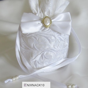 May include: White satin drawstring bag with a white satin bow and a gold and pearl button. The bag has a white floral pattern.