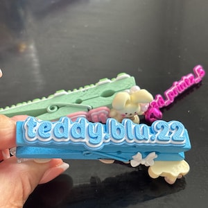 May include: A bright blue clothespin featuring the text "teddy.blu.22" in raised white lettering on the front. The clip is decorated with a small white bow accent on the side and a miniature cream-colored rabbit figure attached to the top surface.