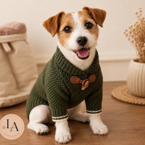 Puede incluir: Un perro pequeño de pelaje blanco y marrón lleva puesto un suéter de punto color verde oliva oscuro. El suéter cuenta con un cuello acanalado, mangas largas con puños a rayas blancas y verdes, y un cierre de palanca marrón con un botón de madera.