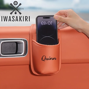 以下が含まれることがあります： オレンジ色のレザー製カードア収納ポケット。黒い筆記体で「Quinn」という名前がプリントされています。ポケットはオレンジ色の内装パネルに取り付けられており、隣にはシルバーのドアハンドルがあります。ポケットには黒いスマートフォンが差し込まれています。